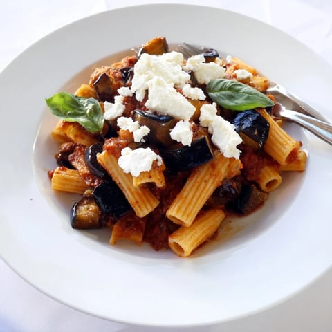 A close-up of rigatoni pasta generously coated in a vibrant red tomato sauce, topped with tender roasted eggplant cubes, fresh basil leaves, and a snowy sprinkle of ricotta salata.
